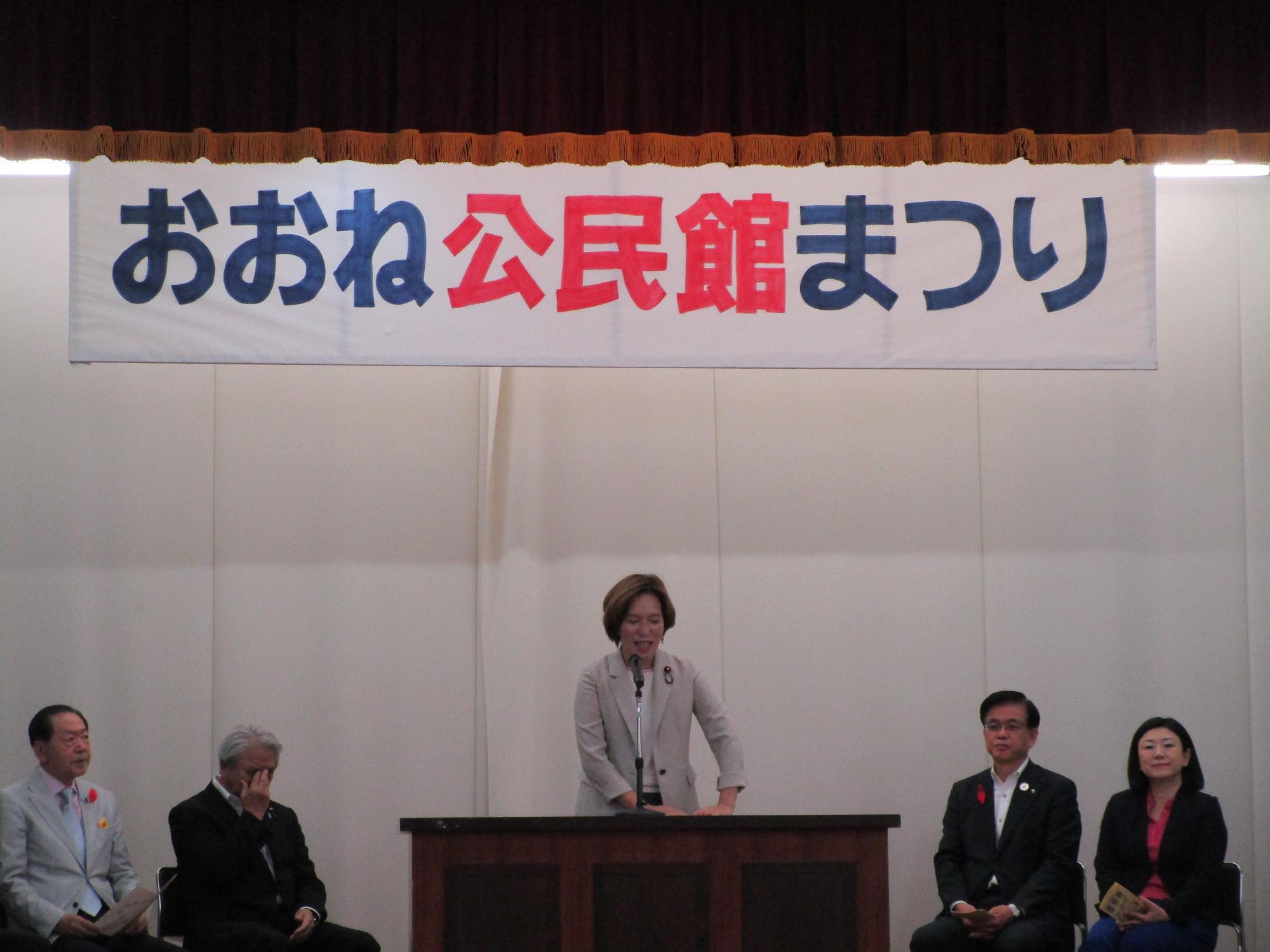 ステージ上の演台に立つベージュのジャケットを着た女性がマイクで話しており、「おおね公民館まつり」と書かれた横断幕の下で数名の来賓らしき男女が着席している様子を捉えた写真
