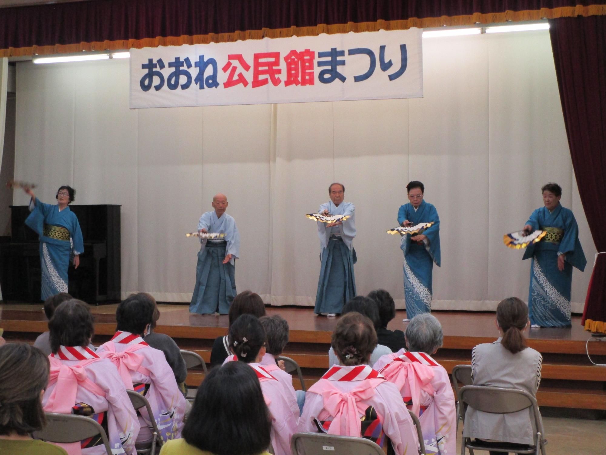 横断幕が掲げられたステージで、扇子を持った着物姿の5人の男女が踊りを披露しており、聴衆も一部着物や浴衣を着て鑑賞している写真