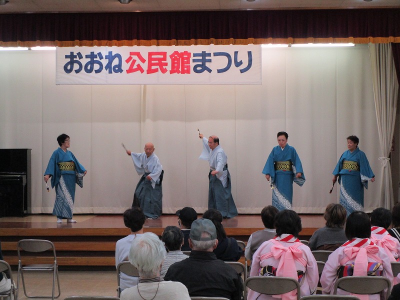 「おおね公民館まつり」と書かれた横断幕の下、ステージ上で男女5人が鮮やかな水色の着物を着て扇子を持ち、観客を前にして踊りを披露している写真