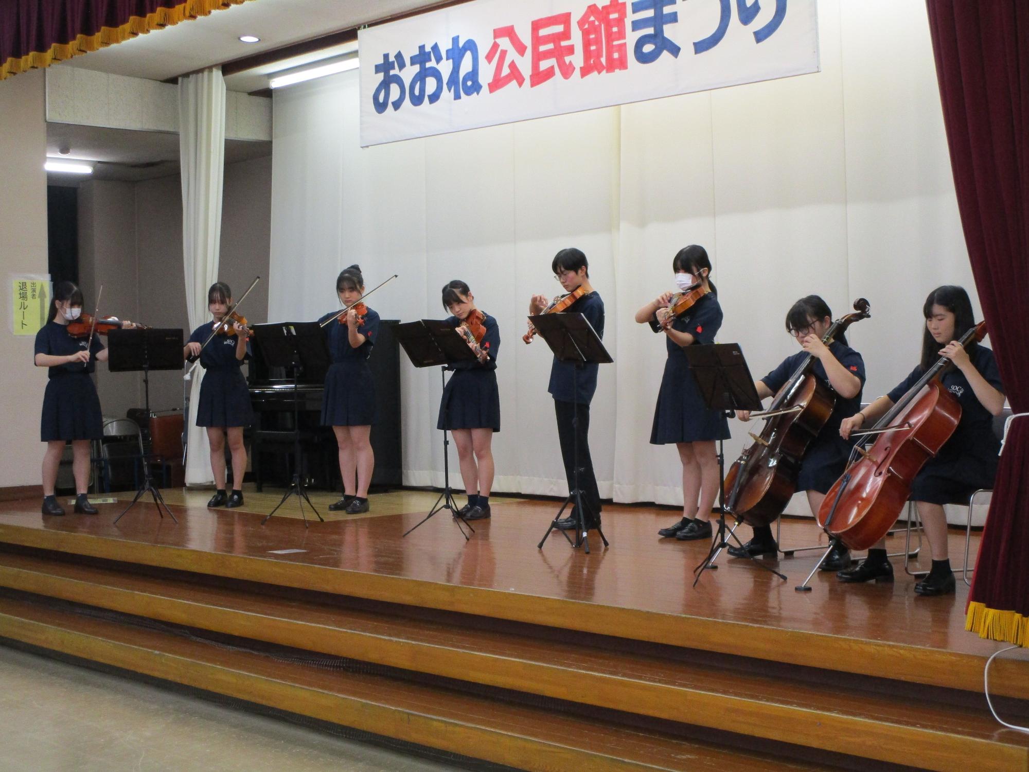 「おおね公民館まつり」と書かれた横断幕が掲げられたステージ上で、お揃いの紺色のティシャツと制服を着用し高校生8人が、譜面台の前に立ちまたは座り、ヴァイオリンとチェロを演奏している写真