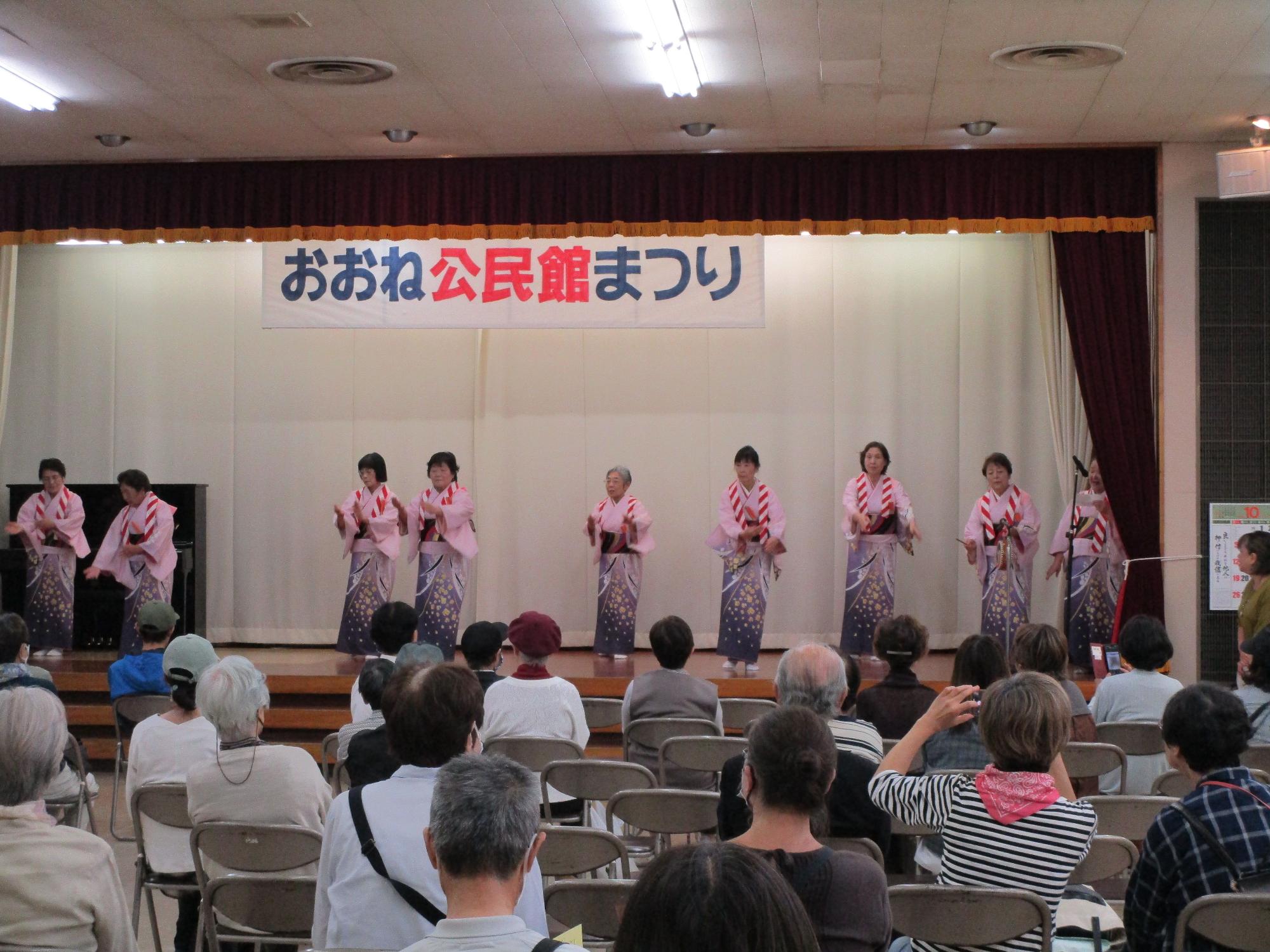 「おおね公民館まつり」と書かれた横断幕が掲げられたステージで、ピンクの着物を着た女性8人が、扇子を手に持ち踊りを披露し右側で1人の女性がマイクで歌を歌っている写真