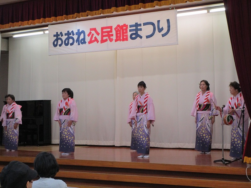 「おおね公民館まつり」と書かれた横断幕が掲げられたステージ上で、ピンクの着物を着た女性7人が踊りを披露し、右側の1人の女性が太鼓を手に持ち叩いている写真