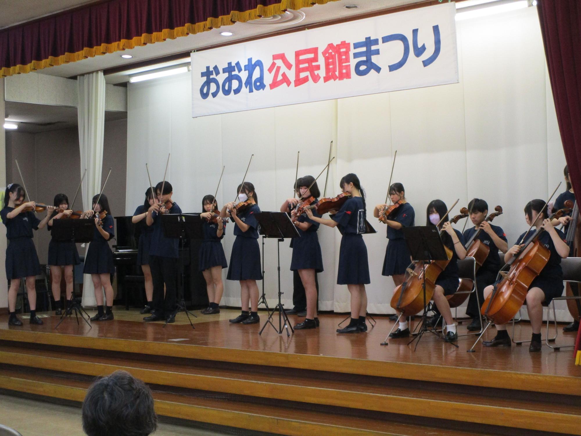 「おおね公民館まつり」の横断幕が掲げられたステージで、お揃いの紺色のティシャツと制服を着用した高校生15名ほどが、ヴァイオリンとチェロなどの楽器で演奏している写真