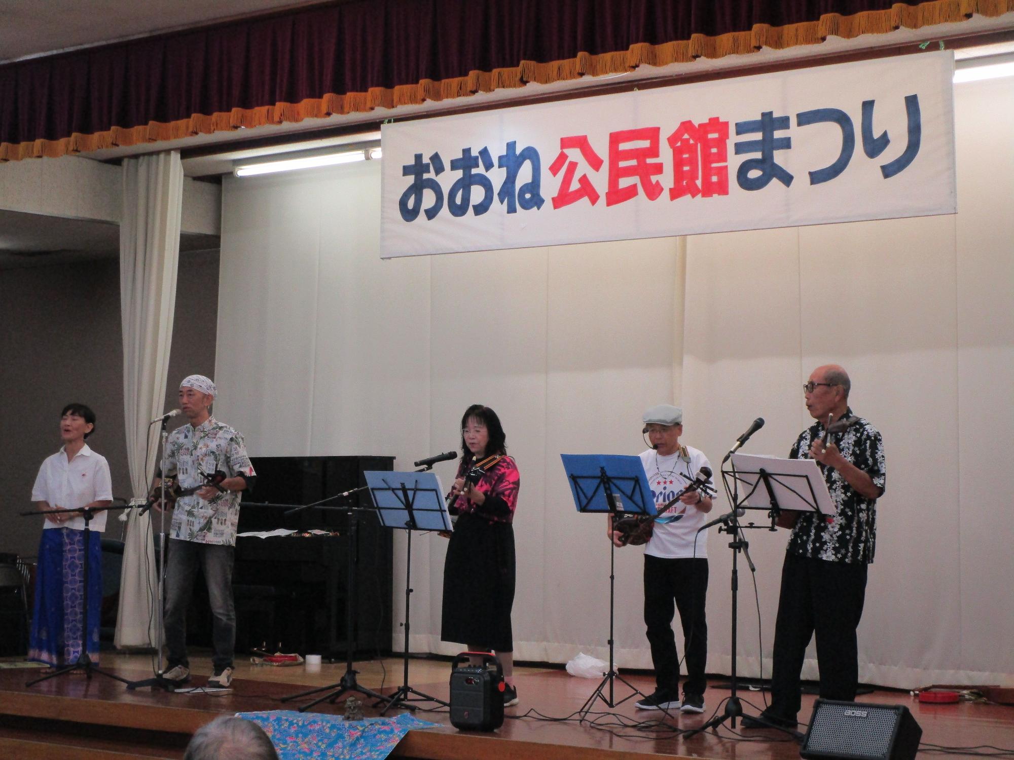 「おおね公民館まつり」と書かれた横断幕が掲げられたステージ上で、男女5人がウクレレのような楽器を演奏したりマイクを持って歌ったりしている様子の写真