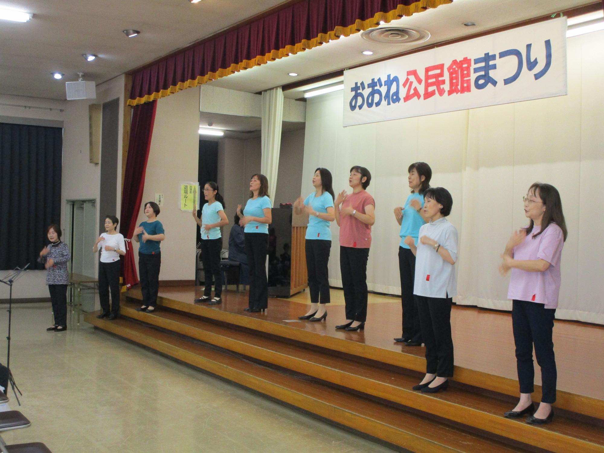 「おおね公民館まつり」と書かれた横断幕が掲げられたステージ上で、水色やピンクなどのTシャツと黒いパンツを着用した女性10人が、観客を前にして身振り手振りで歌を披露している様子の写真