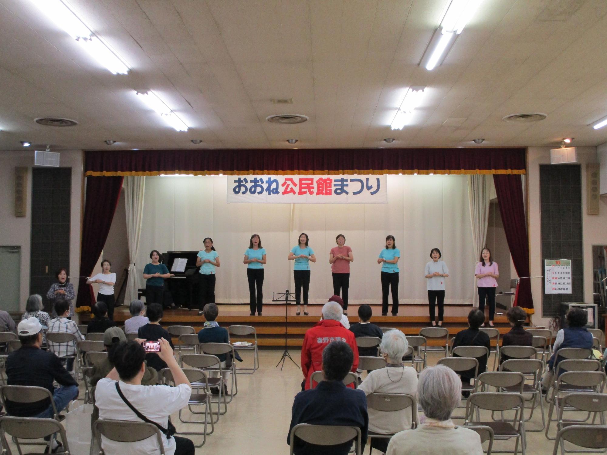 「おおね公民館まつり」と書かれた横断幕が掲げられたステージ上で、女性10人が色違いのTシャツと黒いパンツを着用し、観客を前にして身振り手振りで合唱を披露している様子の写真