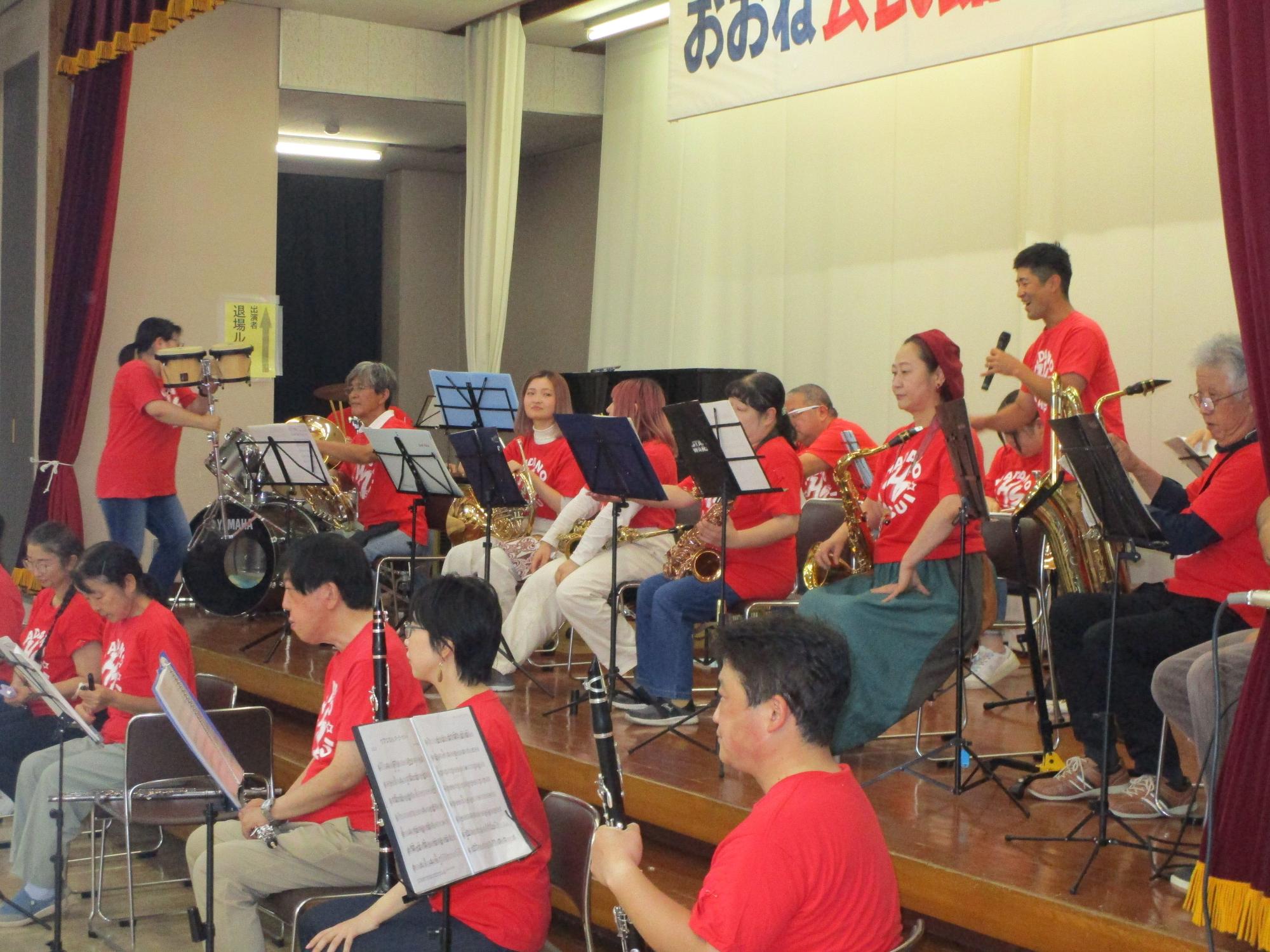 赤色のTシャツを着用した大勢のバンドメンバーが、ステージ上でドラムセット、サックス、クラリネット、ホルンなどの管楽器や打楽器を演奏している、公民館での吹奏楽演奏の写真