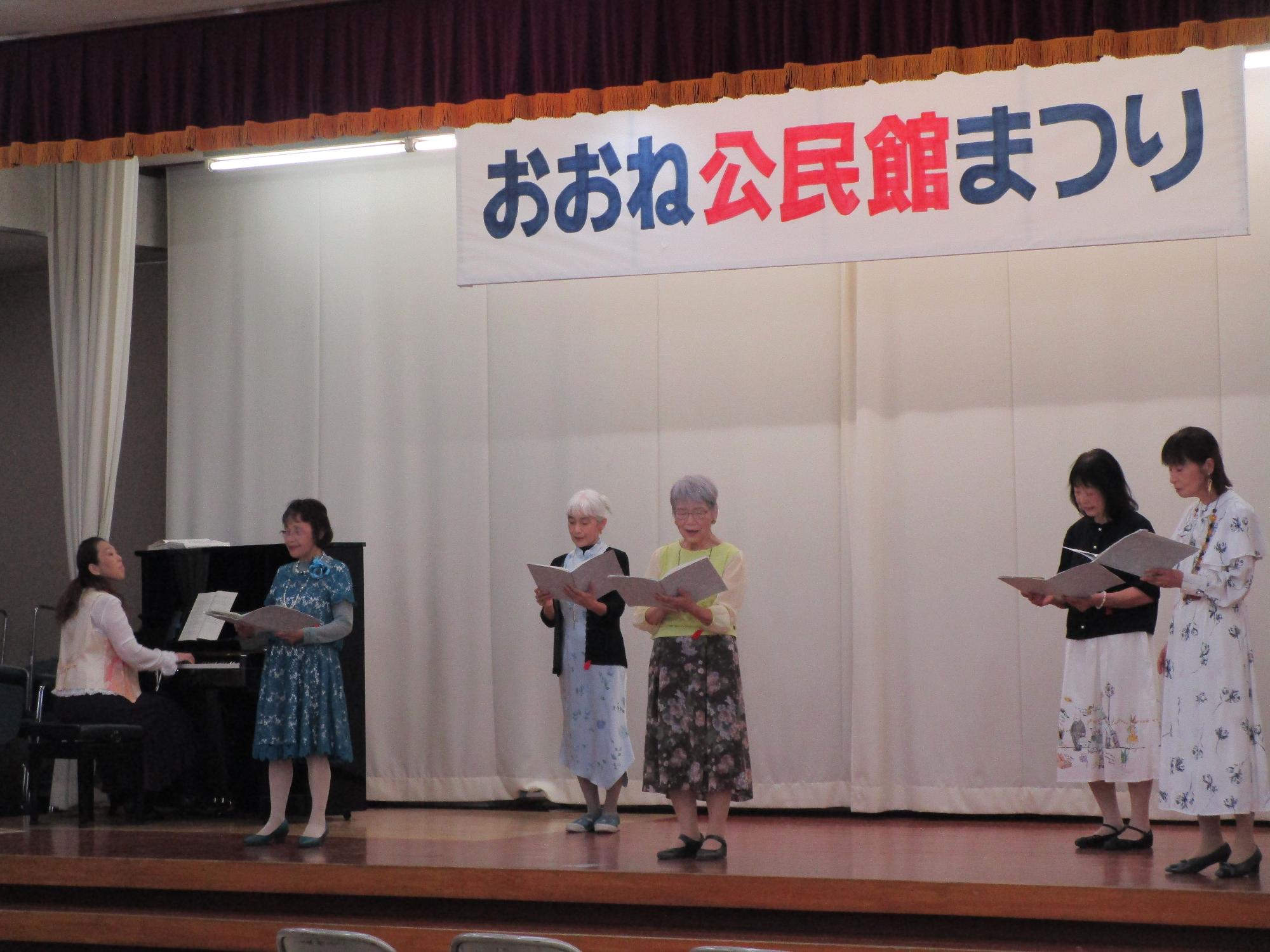「おおね公民館まつり」と書かれた横断幕が掲げられたステージ上で、5人の女性たちがピアノ伴奏に合わせて楽譜を見ながら合唱をしている様子の写真