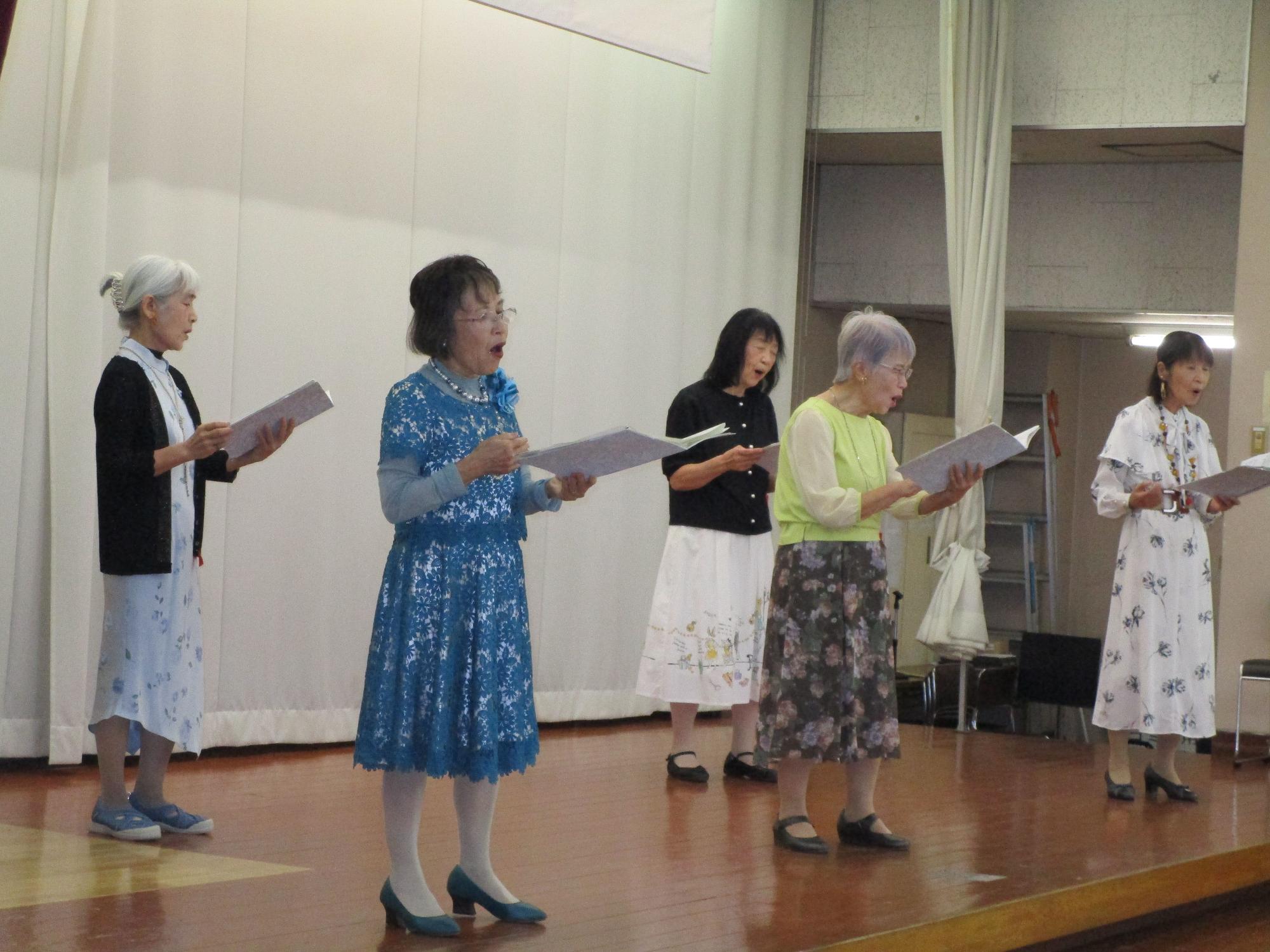 ステージ上で女性5人が、それぞれが異なる華やかなワンピースやスカートを着用し、楽譜を両手に持って合唱を披露している様子の写真