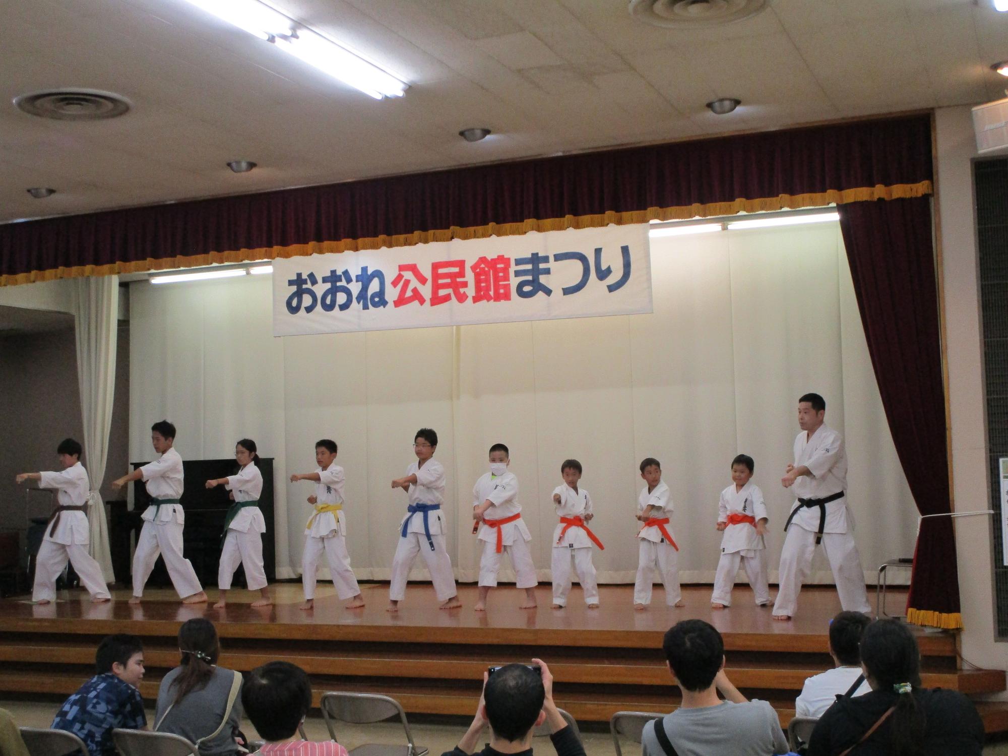 「おおね公民館まつり」の横断幕が掲げられたステージ上で、空手着を着用し帯の色が異なる10人ほどの大人と子供たちが、観客の前で空手の演武を披露している写真