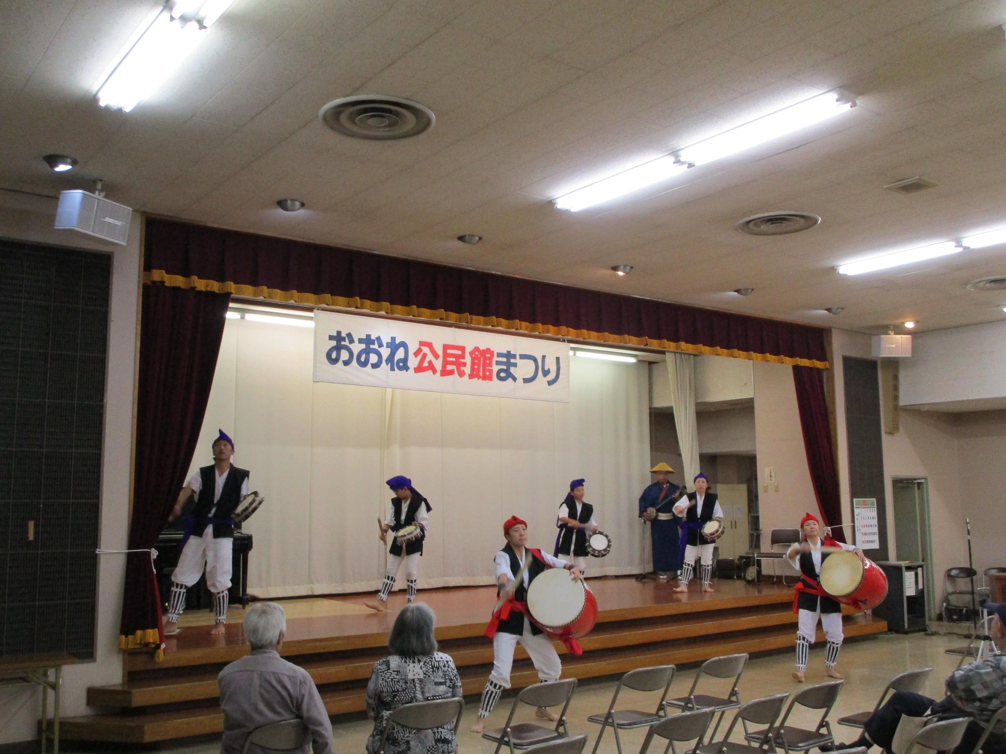 「おおね公民館まつり」と書かれた横断幕が掲げられたステージ上で、赤と青の衣装に身を包んだ男女6人が、大太鼓と小太鼓を力強く演奏して踊っている写真