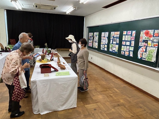 多くの人々が、白い布をかけた長机の上に展示された木彫りなどの作品を鑑賞しており、背景の黒板には植物や食べ物のイラストが多数貼られている、展示会場の様子の写真