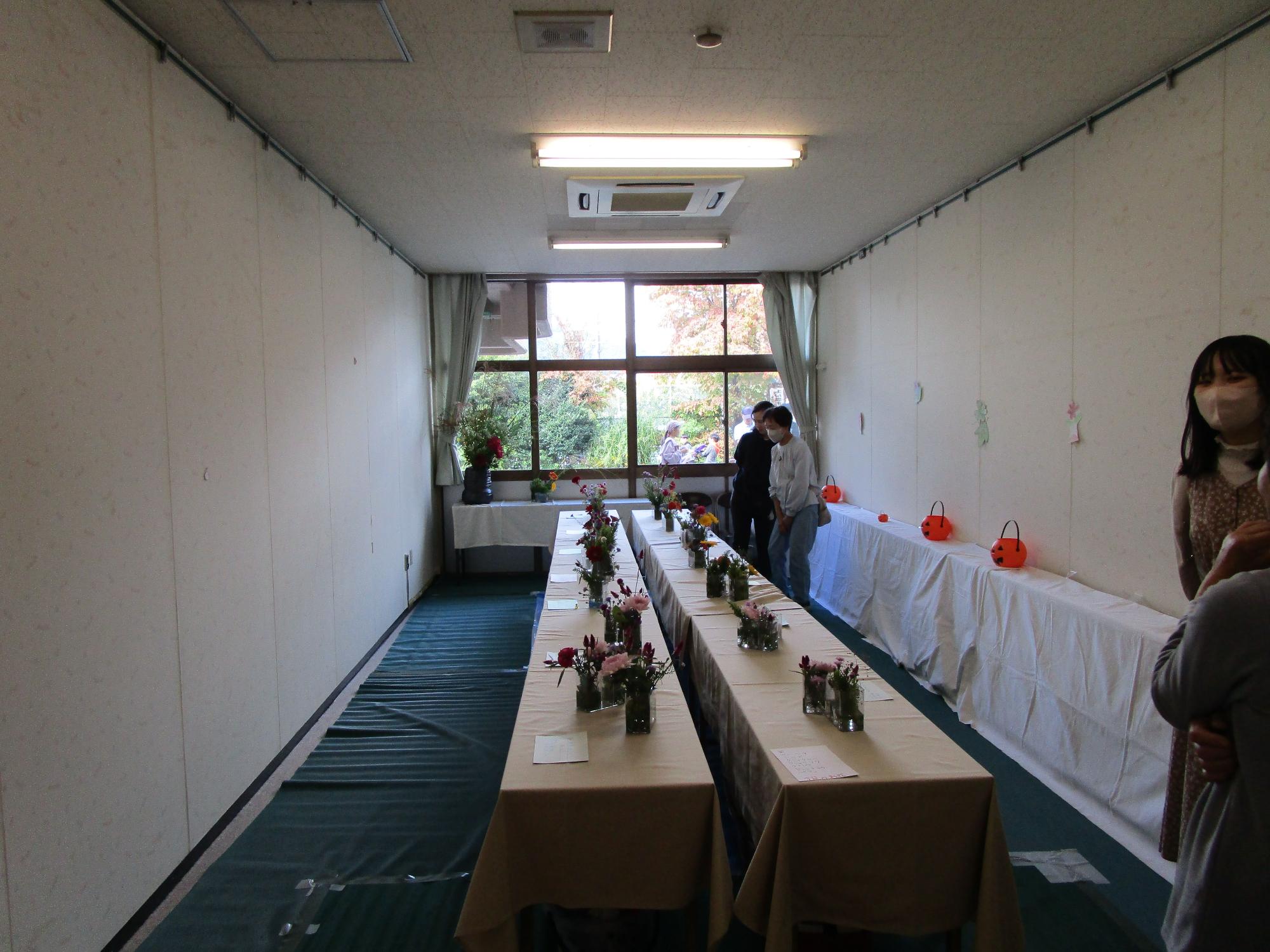 ベージュ色の布がかけられた長テーブルが2列に並び、小さな花瓶に生けられた花々が飾られ、数人の来場者が鑑賞している様子の写真