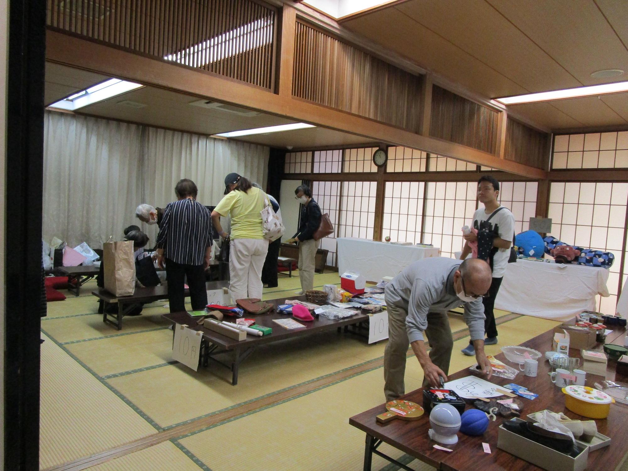 和風の建物内の広間で、畳の上に木製の長い座卓が複数並べられ、多くの人々が集まって品物を見ているバザーの写真