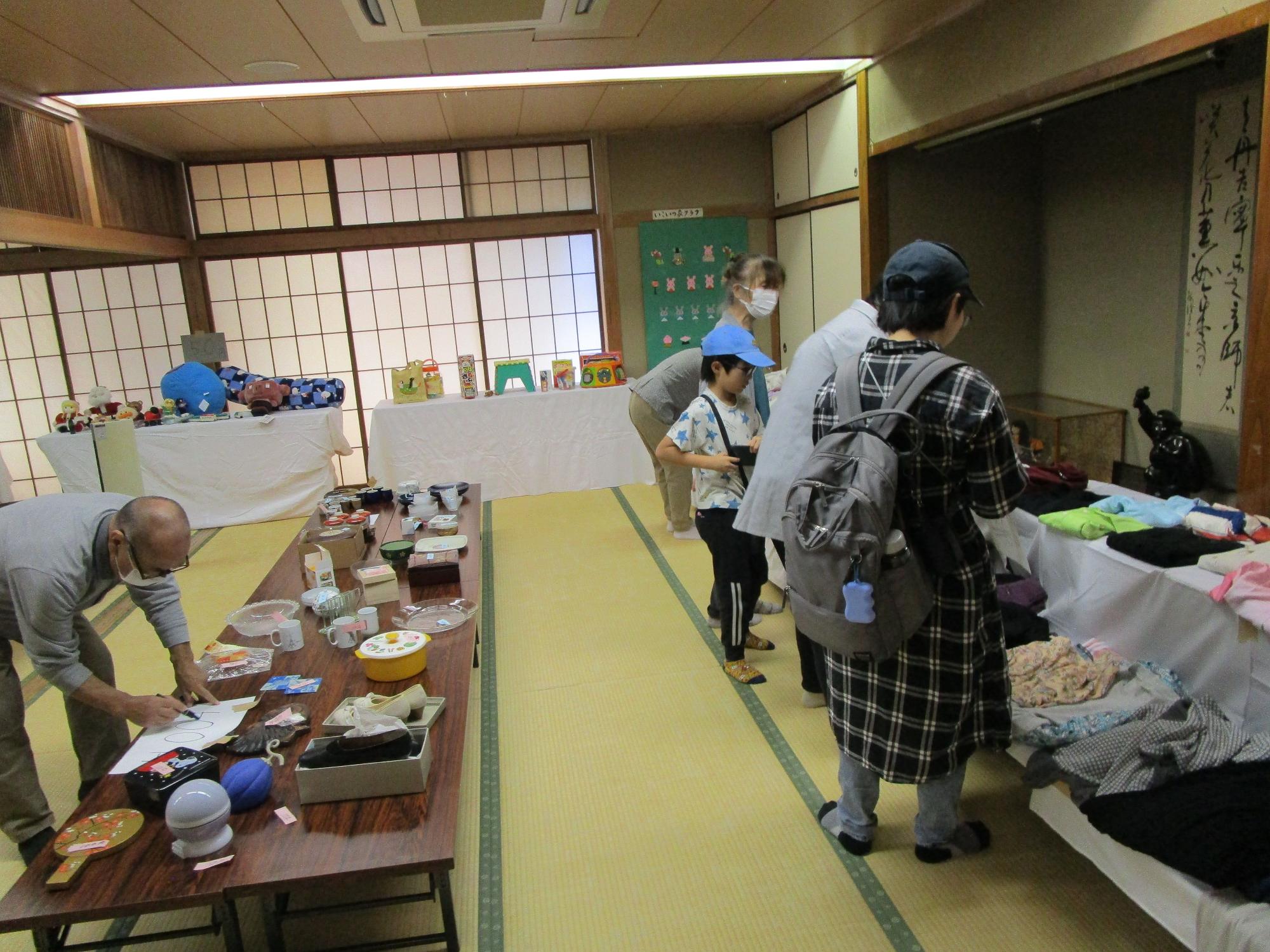 和室の広間の中央に長い座卓が置かれ、ガラス器や雑貨などの物品が並べられ、白い布をかけた長机の上にも服などが展示されており、来場者が熱心に品物を見ているバザーの光景の写真