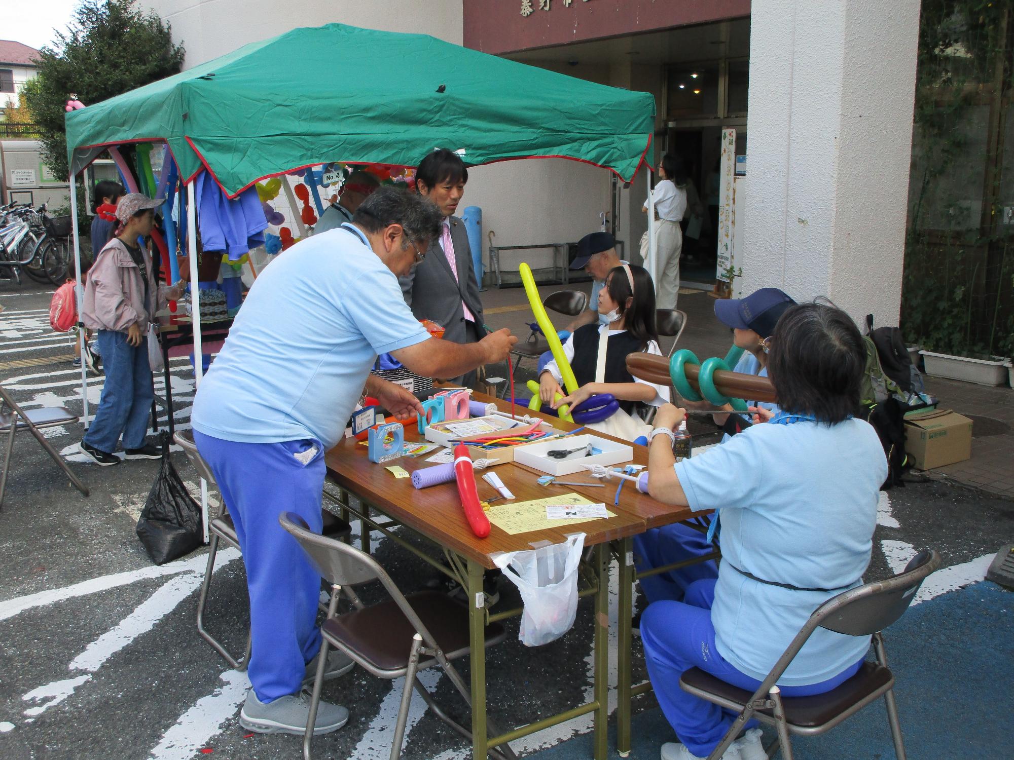 建物の屋外広場に設置された緑色のテントの下で、水色のポロシャツを着た数名のスタッフが、テーブルを囲み風船で作った作品を準備している様子の写真