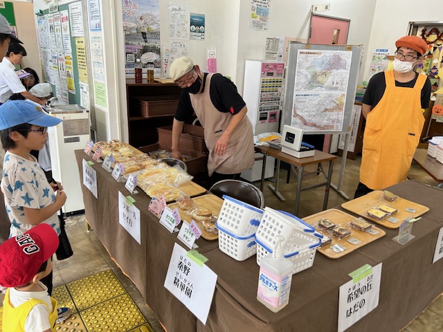 「希望の丘 はだの」と書かれた札のあるブースで、エプロンを着用した2人の男性がパンや焼き菓子を販売しており、数人の子供たちが商品を見てる様子の写真