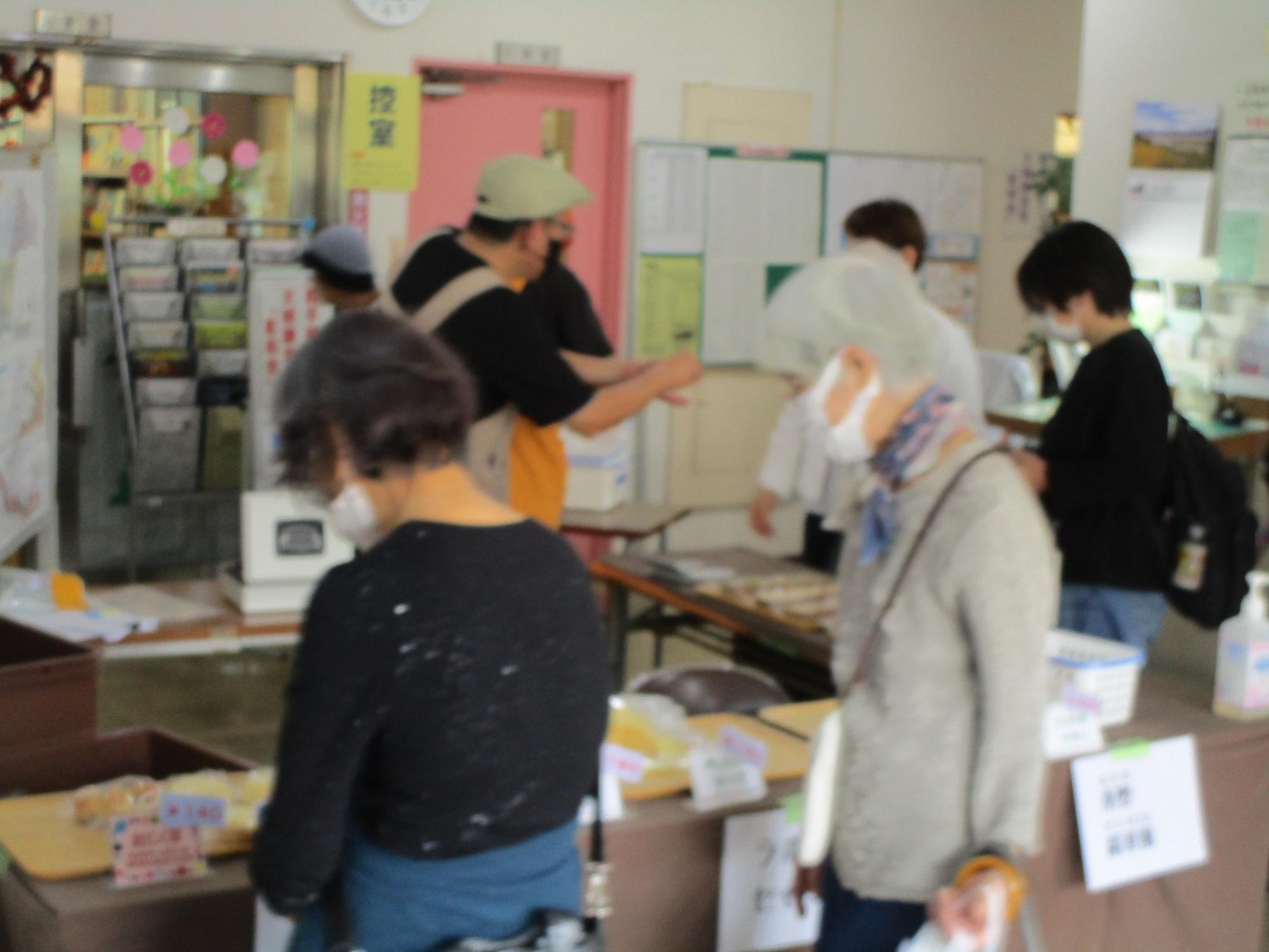 室内で多くの人々がマスクを着用して集まっており、手前のテーブルではパンなどの物品が販売されている様子の写真