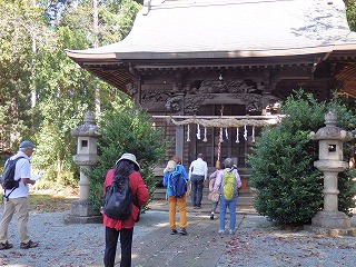 木々に囲まれた境内にある木造の神社拝殿に向かって、リュックを背負った数人の参拝客が、両脇の石灯籠の間を歩いていく後姿を捉えた写真