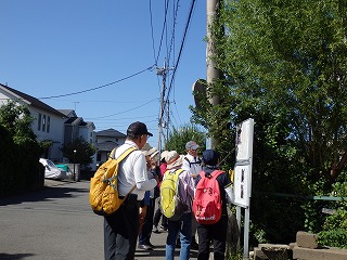 晴天の下、リュックサックを背負い帽子をかぶった数名の人々が緑の屋根瓦と木製の塀を持つ古い寺院のような山門を眺めている様子の写真