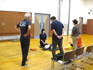 体育館のような広い部屋で、黒いTシャツにSAFETY LEADERと書かれた講師と参加者数名が、床に置かれた成人用と小児用の心肺蘇生訓練用人形を用いて実習を受けている様子の写真
