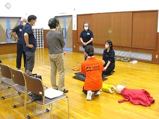 フローリングの体育館のような部屋で、オレンジ色のTシャツにSAFETY LEADERと書かれた指導者と数名の参加者が、床に置かれた乳児用と成人用の訓練用人形を用いて心肺蘇生法の指導を受けている様子の写真