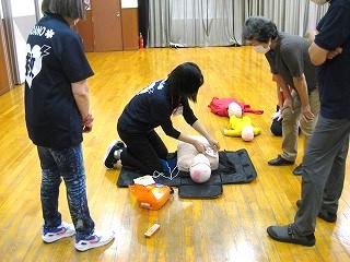 体育館のようなフローリングの部屋で、指導員と思われる女性が成人用の心肺蘇生訓練用人形にAEDのパッドを装着し、数名の参加者がそれを取り囲んで実習を見学している様子の写真