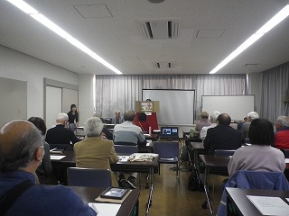 白いスクリーンとプロジェクターが設置された教室で、机と椅子に座った多くの参加者が、前方に立つ講演者の方を向いて熱心に耳を傾けている様子の写真