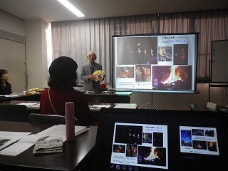 教室で行われているセミナーで、講師が何かを手に持ちながら、スクリーンに画像が映し出されているスライドを見せ、参加者に向かって話している様子の写真