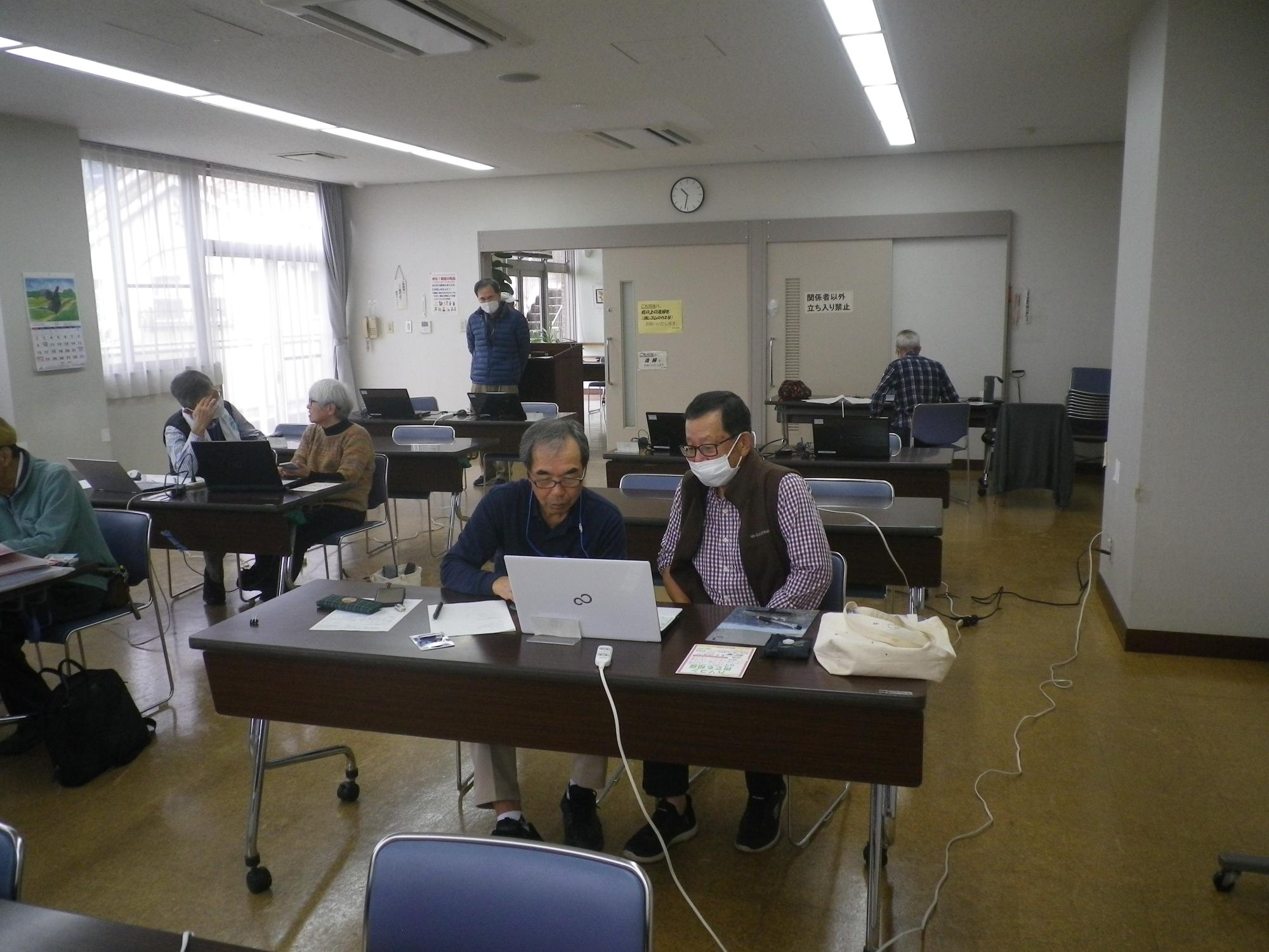 明るい教室で、マスクを着用した参加者たちが向かい合ってパソコンの画面を見たり、各自の机で作業をしたりしている、パソコン教室の様子の写真