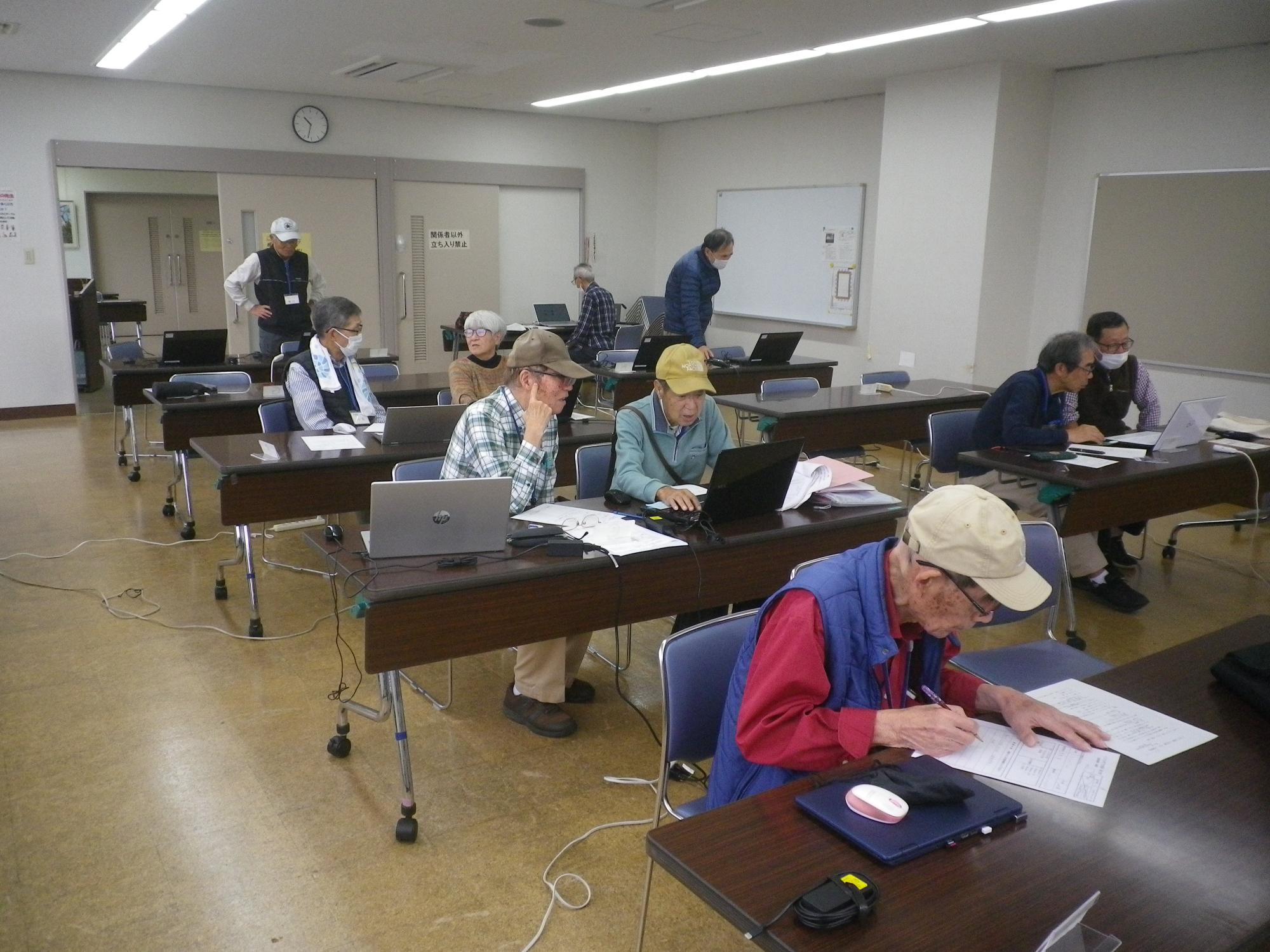 教室で数人の参加者たちが、各自の前に置かれたノートパソコンに向き合ったり、配布された資料に書き込んだりして、パソコン講座に参加している様子の写真