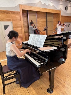 木目調の壁の会場で、黒いグランドピアノを弾くロングドレスの女性と、後方でチェロとヴァイオリンを演奏する二人を捉えた室内楽演奏の写真