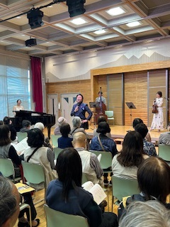 木目調の壁のステージ前で、青いスーツの男性ボーカリストがマイクでを持ち、その横にはグランドピアノを弾く女性、コントラバス、ヴァイオリン奏者が立ち、椅子に座って楽譜のようなものを持つ多くの参加者がステージを見ている様子の写真