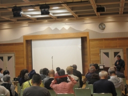 講堂のような会場で、聴衆が正面の巨大な白スクリーンと、スクリーン右側で立って話す男性の話を聞いている様子の参加者を後ろから写した写真