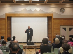 講堂のような会場で、参加者が正面の巨大な白スクリーンと、スクリーンの前で立って話す男性の話を参加者が聞いている写真