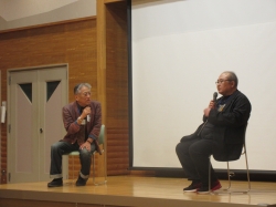 木目調の壁のステージ上で男性二人が椅子に座りマイクを持ち話しているのを参加者が聞いている写真