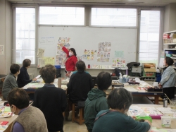 教室で、一人の女性講師がホワイトボードに貼られたカラフルな作品を指し示しながら、集まった数名の生徒に向けて指導している様子を捉えた写真