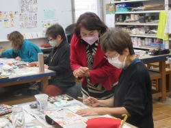 赤いジャケットを着た講師が、制作中の生徒に寄り添い、真剣な表情で手元の作品についてアドバイスしている様子の写真