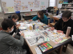 四人の生徒がテーブルを囲んで座り、それぞれ水彩絵の具のパレットや筆を使いながら、テーブルに広げたカラフルな絵手紙の制作に熱心に取り組んでいる写真