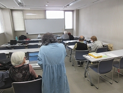 白い壁と大きな窓のある講義室で、数名の高齢者が机に向かってノートパソコンを開き、立っている講師らしき人物に教えを受けながら学習している様子の写真