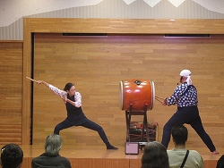 木目調のステージ上で、2人の演奏者が1つの大きな和太鼓を両側から力強く打ち鳴らし、観客に向けて躍動的なパフォーマンスを披露している様子の写真