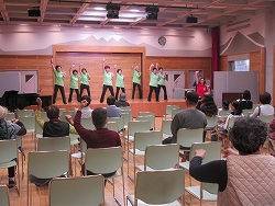 黄緑色のお揃いのTシャツを着た数人の男女が、ステージ上で観客に向けて両手を挙げた統一感のあるダンスを披露している写真