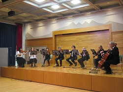 山脈の背景と木目調の壁があるステージ上で、複数の演奏者がバイオリン、ビオラ、チェロなどの弦楽器を演奏し、観客に向けて披露している様子の写真