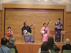 和服姿の4人の演奏者がステージ上で三味線と和太鼓を使い、観客に向けて日本の伝統音楽を披露している様子の写真