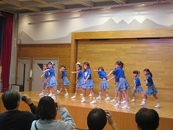 ステージ上青いトップスとフリルのついたデニム風のミニスカートのお揃いの衣装を着た十人ほどの女の子たちがダンスを披露している様子写真