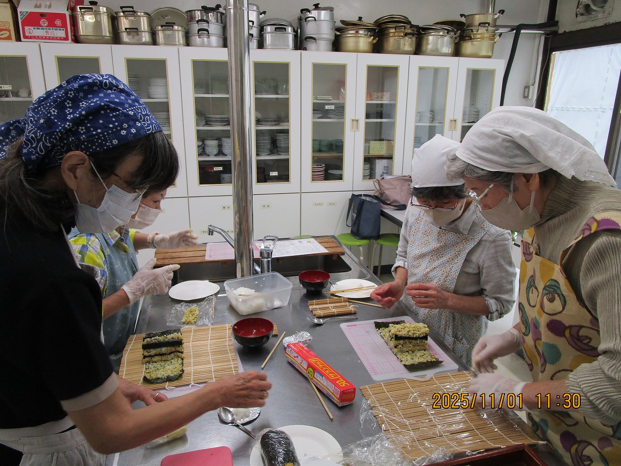 三角巾、マスクを着用した数人の女性たちが、調理台に置かれた巻きすの上で海苔にご飯や具材を広げ、飾り巻き寿司を調理している様子の写真
