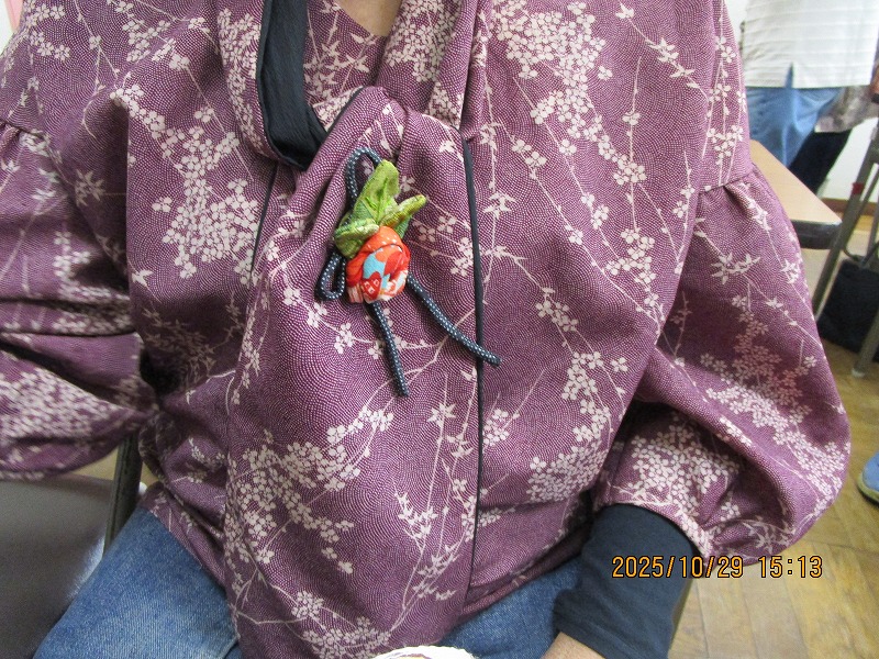 白く細かい花柄が織り込まれた紫色の和風ブラウスを着た人が、黒い紐状のリボンと緑と赤の小さなつまみ細工の飾りを胸元に付けている写真