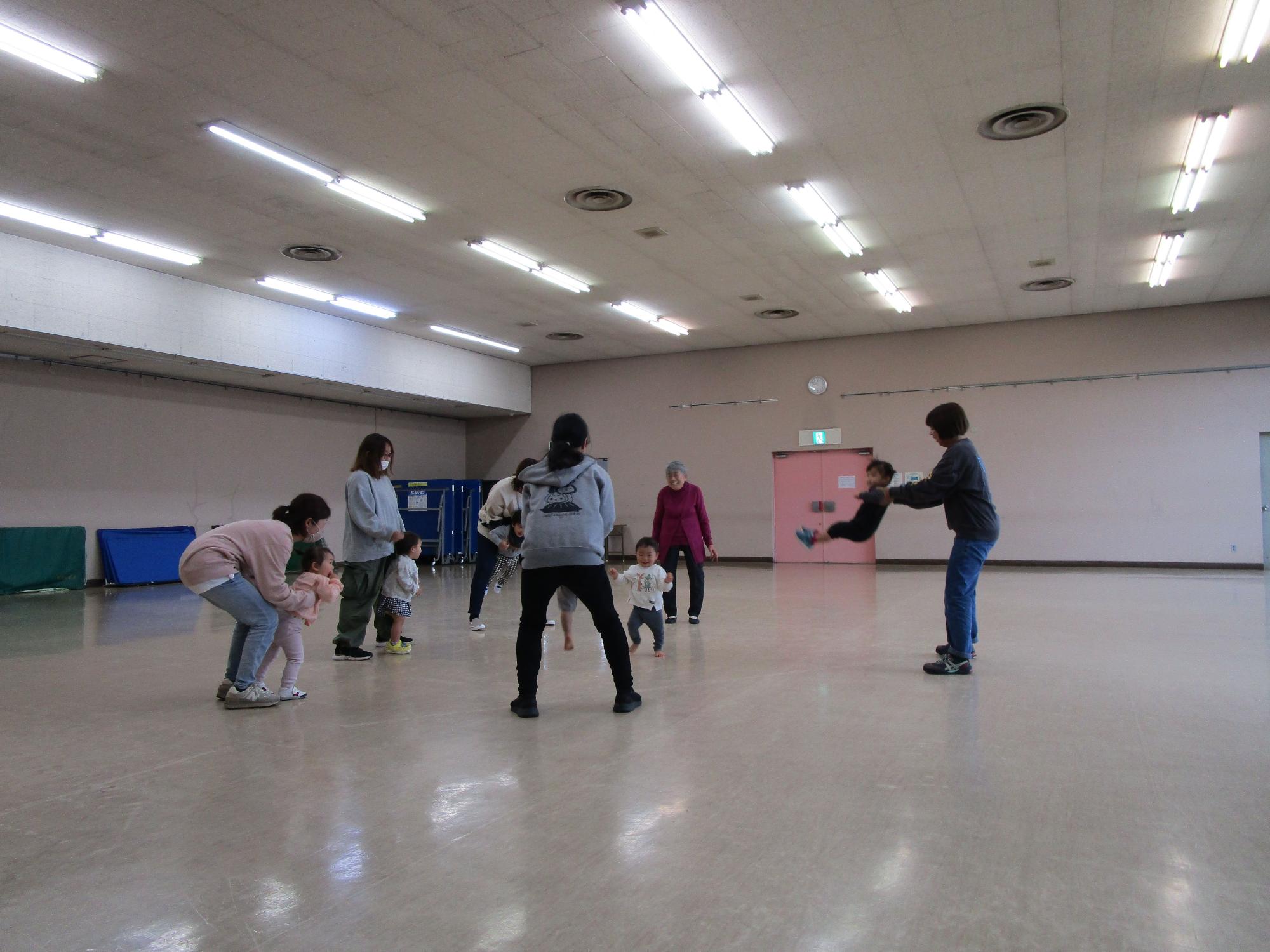 広いホールで、幼児の親子が立ったりしゃがんだりしながら遊んだり抱っこしたりしている写真