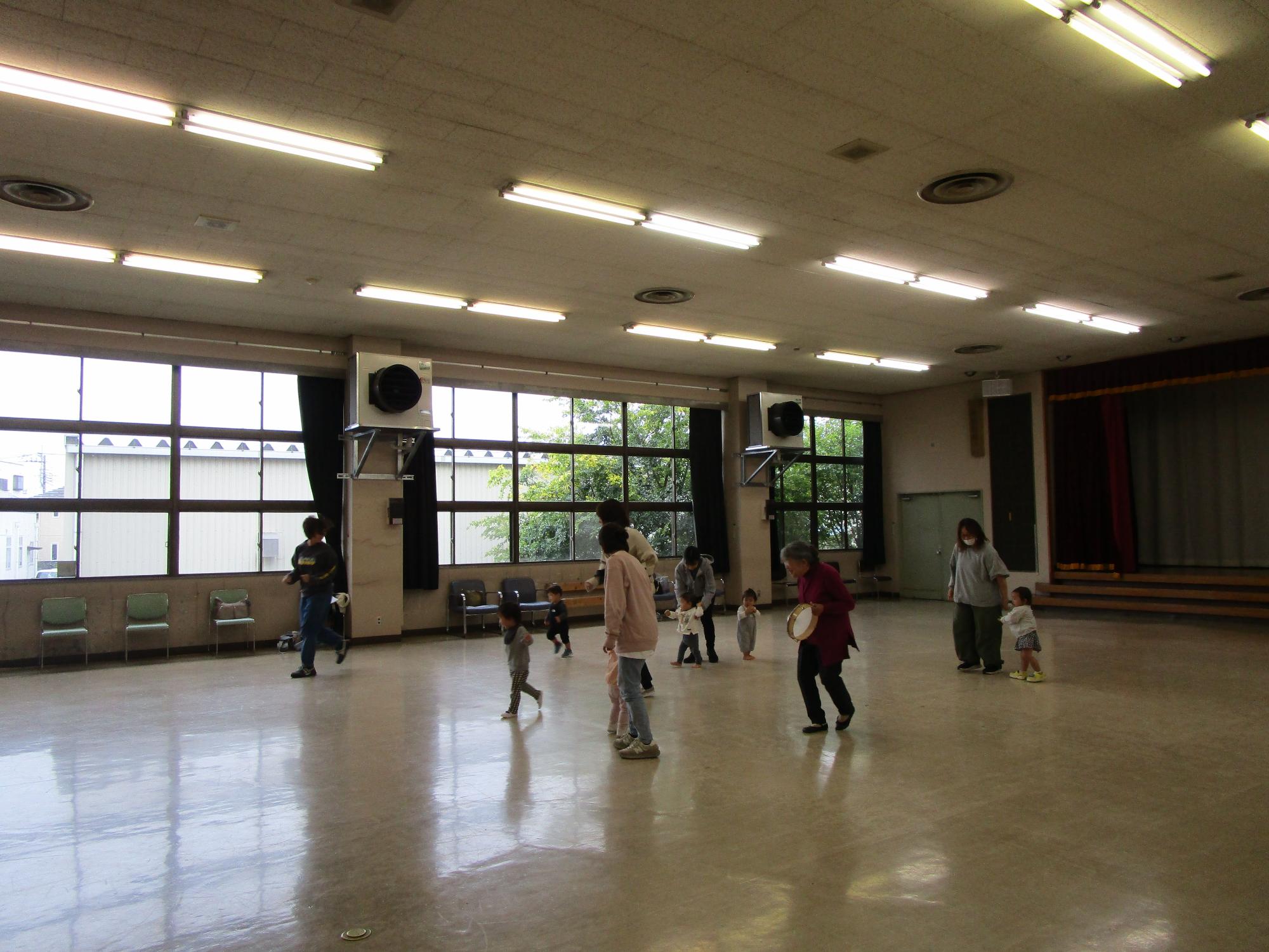 広くて明るいホールで、タンバリンを持った大人たちが幼い子どもたちと一緒に床を走り回っている写真