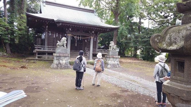 森に囲まれた木造の社殿の前に、左右一対の狛犬が鎮座しており、数人の参拝客が敷石の道を通りながら拝殿に向かっている様子の写真